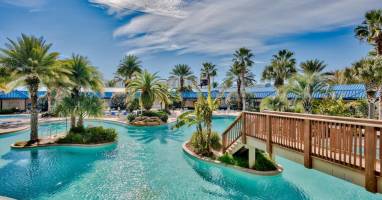 Resort-style pool w/ winding waterways, palm trees, and a sunny bridge connecting lush islands.