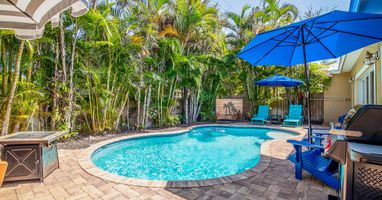 Private freeform heated pool and tropical paver patio at Coastal Cottage, a 3-bedroom vacation home in Holmes Beach on Anna Maria Island. Lush palm landscaping creates a secluded backyard oasis with lounge seating, shaded umbrellas, and a gas grill — perf