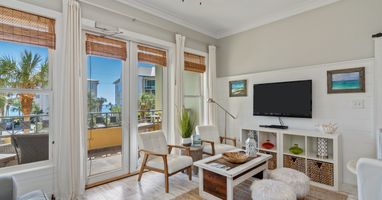 Light-filled living area with coastal flair, featuring comfy seating, a flat-screen TV, and glass doors that open to a palm-lined balcony—perfect for a laid-back 30A getaway.