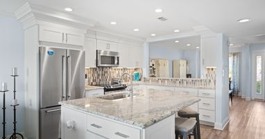 Modern kitchen with a large granite island, stainless steel appliances, white shaker cabinets, tile backsplash, and recessed lighting. Features include bar seating, hardwood floors, and a mirrored wall for added depth.