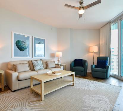 Bright living room w/ cozy seating, coastal artwork, and sliding doors opening to the balcony.