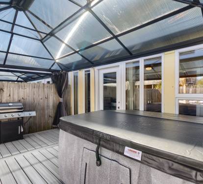 Cozy covered outdoor patio with string lights, a private hot tub, and a gas grill perfect for relaxing evenings and outdoor dining in a fenced backyard space.
