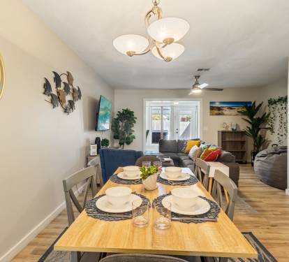 Open-concept dining and living area featuring a cozy coastal-inspired design, with a fully set dining table, comfortable sectional seating, and stylish beach-themed d&eacute;cor. Bright, inviting space perfect for gathering with family and friends.
