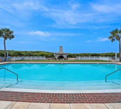 Large resort-style pool surrounded by palm trees and elegant stonework with outdoor fireplace beneath brilliant blue skies.