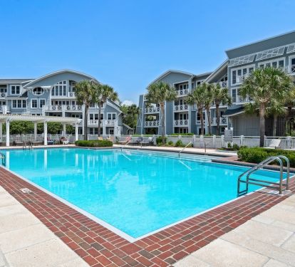 Large swimming pool with brick border surrounded by multi-story resort buildings and tropical palm trees under clear blue skies.