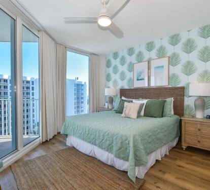 Calm bedroom w/ a bed, leafy accent wall, and large windows bringing in natural light.