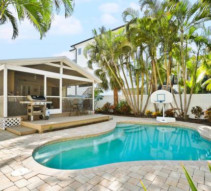 This tropical backyard features a private swimming pool, a built-in grill, and lush palm landscaping—perfect for relaxing outdoor meals after a beach day.