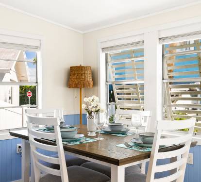 This bright dining nook features large windows, coastal blue accents, and a wooden table set for four—perfect for casual meals in a beach cottage setting.