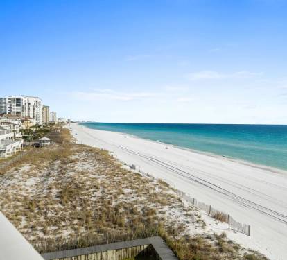 Pristine white sand beach stretches along turquoise waters with coastal buildings and natural dune vegetation.