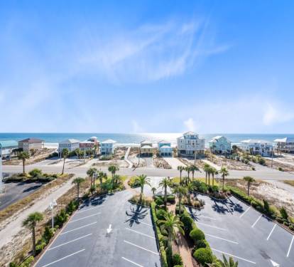 Aerial view of beachfront community with colorful vacation homes and landscaped parking area surrounded by palm trees and white sand dunes.