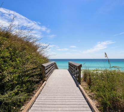 Wooden boardwalk leads through coastal vegetation to pristine turquoise waters and sandy beach under brilliant blue skies.