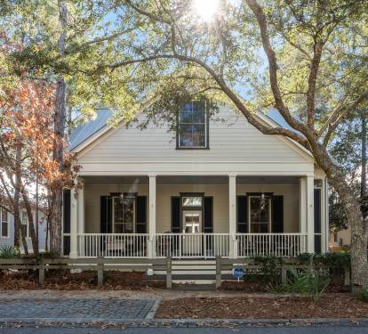 A charming white cottage with wraparound porch nestled among mature oak trees on a quiet cobblestone street.