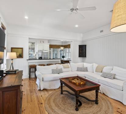 Sink into plush white sofas in your bright living room, where coastal charm meets modern comfort with soaring ceilings and open kitchen views.