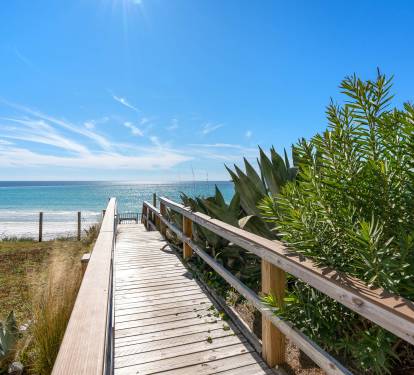 A scenic wooden boardwalk leads through coastal vegetation to pristine sandy shores and crystal-clear waters.