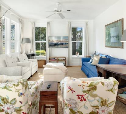 Your bright living room invites relaxation with cozy seating, coastal artwork, and natural light streaming through windows overlooking lush greenery.