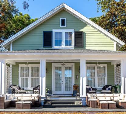 Charming green cottage with wraparound porch featuring comfortable outdoor seating perfect for morning coffee or evening relaxation.