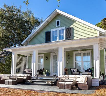 Charming green cottage with spacious wraparound porch featuring comfortable outdoor seating surrounded by mature trees on sunny day.