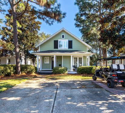 Charming green cottage with front porch and tree-lined driveway, featuring convenient parking and peaceful neighborhood setting.