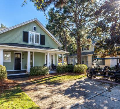 Charming green cottage with front porch and convenient parking area, nestled among mature trees in a peaceful neighborhood setting.