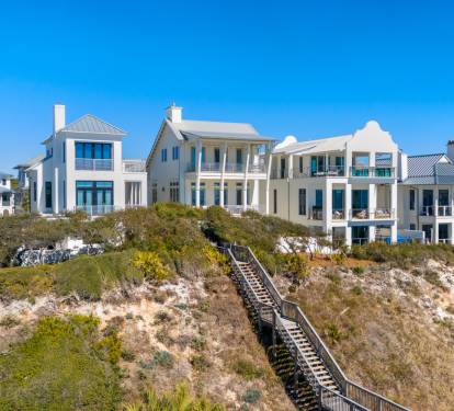 Stunning beachfront homes line the coast with natural dune vegetation and wooden boardwalk access under brilliant blue skies.