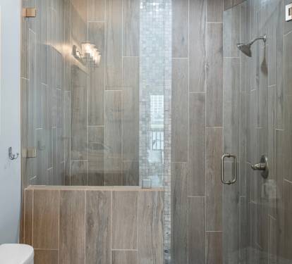 The en-suite bathroom features a luxurious walk-in shower with sleek wood-look tile, mosaic accent strip, and glass doors—blending spa-like comfort with modern coastal design.