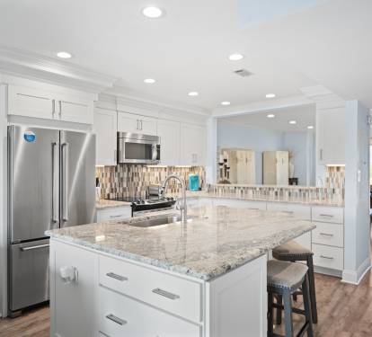 Modern kitchen with a large granite island, stainless steel appliances, white shaker cabinets, tile backsplash, and recessed lighting. Features include bar seating, hardwood floors, and a mirrored wall for added depth.