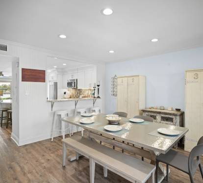 Bright dining area with seating for six, barstools at the kitchen counter, and LVP wood floors. Features a clean coastal style with light furnishings and an open layout.