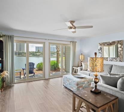 Bright, lakefront living room with large glass doors, a cozy sectional, stylish decor, and serene water views. Features patio access and warm LVP wood flooring for a relaxed, coastal feel.