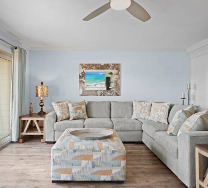 Cozy living room with a large sectional sofa, patterned ottoman, warm LVP wood floors, and beach-themed decor. Soft tones and natural light create a relaxed, inviting atmosphere.