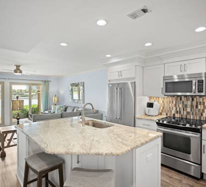 Open-concept kitchen with granite island, stainless steel appliances, and modern backsplash. Flows into a bright living area with lake views and stylish, coastal decor.
