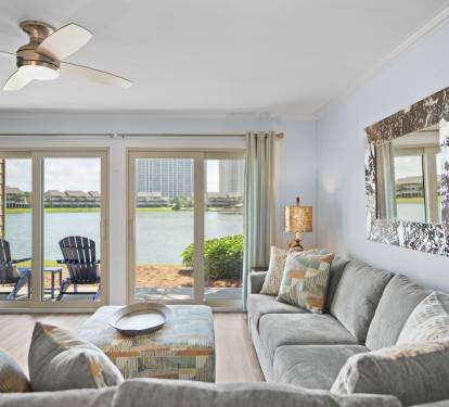 Coastal living room with plush sectional, patterned ottoman, and large glass doors opening to a lakefront patio. Light-filled and serene with stylish, beachy accents.