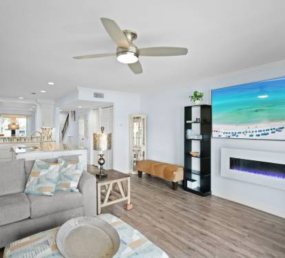 Open, coastal-style living space with LVP wood floors, large screen tv, modern electric fireplace, and beachy decor. Features comfy seating, ceiling fan, and a seamless flow into the dining and kitchen areas.