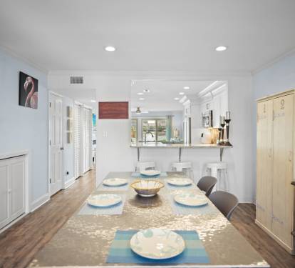 Bright dining area with a modern coastal feel, featuring a large table set for six, bar seating at the kitchen pass-through, and warm wood-look flooring. Open layout connects seamlessly to the kitchen and living spaces.