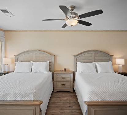Guest bedroom with two queen beds, matching wood headboards and nightstand, crisp white bedding, and a ceiling fan. Neutral tones and wood floors create a calm, inviting atmosphere.