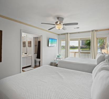 Lakeview guest bedroom with two queen beds, ensuite bathroom, wall-mounted TV, and sliding doors to a private balcony. Features white bedding, soft natural light, and a ceiling fan for comfort.