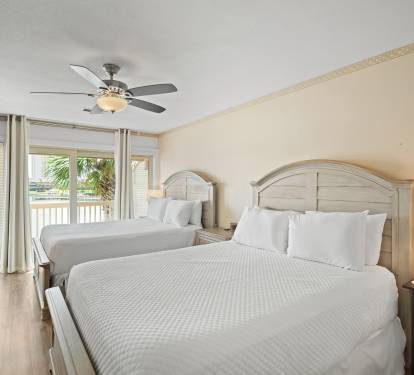 Bright guest bedroom with two queen beds, crisp white bedding, and light wood furniture. Features a ceiling fan, warm neutral walls, and access to a balcony with lake views.