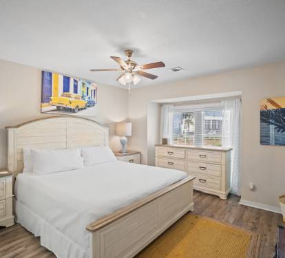 Inviting primary bedroom with a king bed, light wood furniture, and bright window nook. Features soft neutral tones, wood-look flooring, a ceiling fan, and vibrant wall art for a cozy, beachy feel.