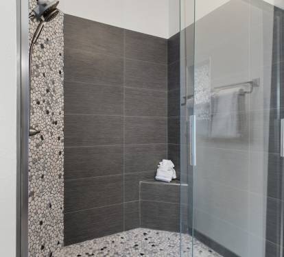 Step into the spa-like walk-in shower with sleek dark tile, pebble flooring, a built-in bench, and glass doors.