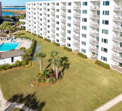 Aerial view of white oceanfront buildings with landscaped lawns, palm trees, and swimming pool, with water visible in the distance.