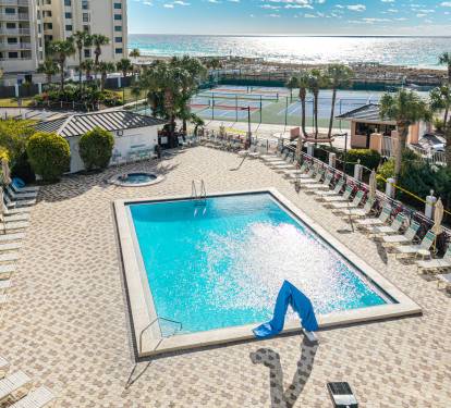 Beachfront resort pool area featuring multiple swimming pools, hot tub, tennis courts, and direct beach access with palm trees.