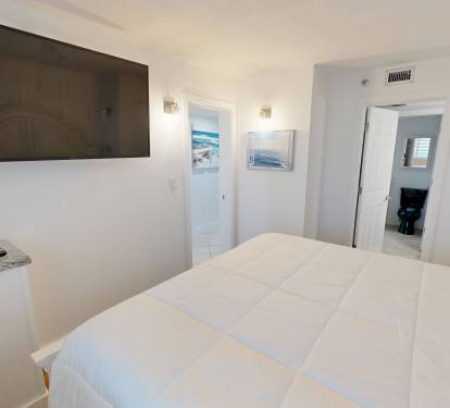 Sink into the plush comfort of your coastal bedroom with crisp white linens, a mounted flat-screen TV, and calming seaside art to set the mood for your perfect beach escape.