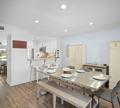 Bright dining area with seating for six, barstools at the kitchen counter, and LVP wood floors. Features a clean coastal style with light furnishings and an open layout.