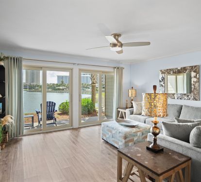 Bright, lakefront living room with large glass doors, a cozy sectional, stylish decor, and serene water views. Features patio access and warm LVP wood flooring for a relaxed, coastal feel.