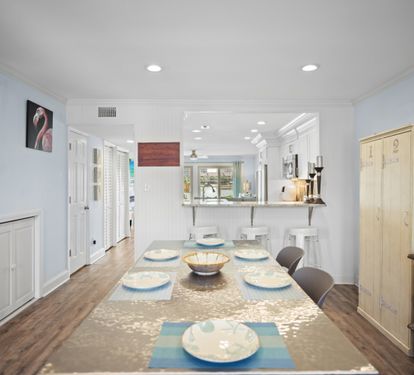 Bright dining area with a modern coastal feel, featuring a large table set for six, bar seating at the kitchen pass-through, and warm wood-look flooring. Open layout connects seamlessly to the kitchen and living spaces.