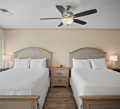 Guest bedroom with two queen beds, matching wood headboards and nightstand, crisp white bedding, and a ceiling fan. Neutral tones and wood floors create a calm, inviting atmosphere.