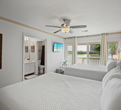 Lakeview guest bedroom with two queen beds, ensuite bathroom, wall-mounted TV, and sliding doors to a private balcony. Features white bedding, soft natural light, and a ceiling fan for comfort.