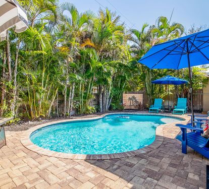Private freeform heated pool and tropical paver patio at Coastal Cottage, a 3-bedroom vacation home in Holmes Beach on Anna Maria Island. Lush palm landscaping creates a secluded backyard oasis with lounge seating, shaded umbrellas, and a gas grill — perf