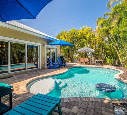 The free-form pool is the centerpiece of Coastal Cottage's private courtyard, framed by a brick sun deck, shaded lounge seating, and lush palm landscaping — a quiet retreat just steps from the back door.