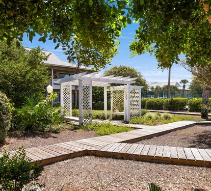 Beautifully landscaped garden with wooden walkways and white pergola structure surrounded by mature trees and tropical plants.