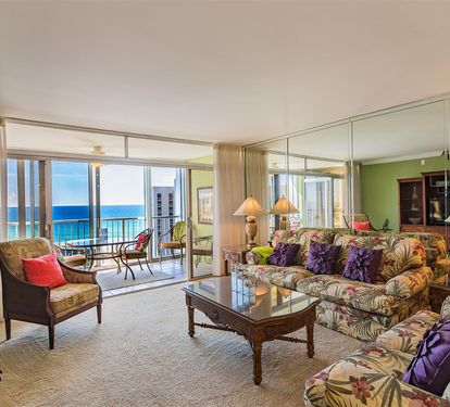 Step into your tropical living room where plush floral seating meets stunning ocean views through floor-to-ceiling windows.